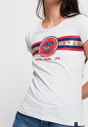 Kobieta w jasnoszarej koszulce z czerwonymi, niebieskimi i złotymi paskami oraz okrągłym logo tygrysa z napisem „Superdry Vintage Tokyo City, Trade Mark JPN”.