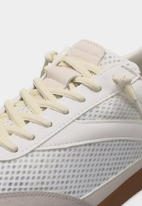 Baskets blanches avec lacets beiges, côtés en mesh, languette rembourrée et semelle marron, montrées en gros plan avec un nœud de lacet attaché.