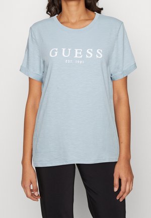 Frau trägt ein hellblaues Kurzarm-T-Shirt mit weißem "GUESS EST. 1981"-Aufdruck, kombiniert mit schwarzen Hosen, steht vor einfachem Hintergrund.