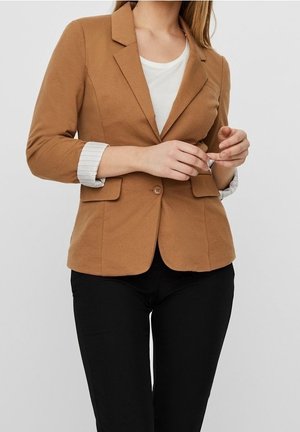 Frau richtet die Manschette eines braunen Blazers mit einem Knopf, trägt darunter ein weißes Top und schwarze Hosen.