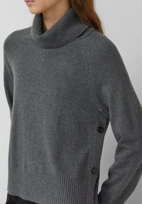 Pull en col roulé gris avec une texture côtelée, doté de boutons sur les côtés et d'une coupe décontractée. Le tissu est doux et légèrement extensible.
