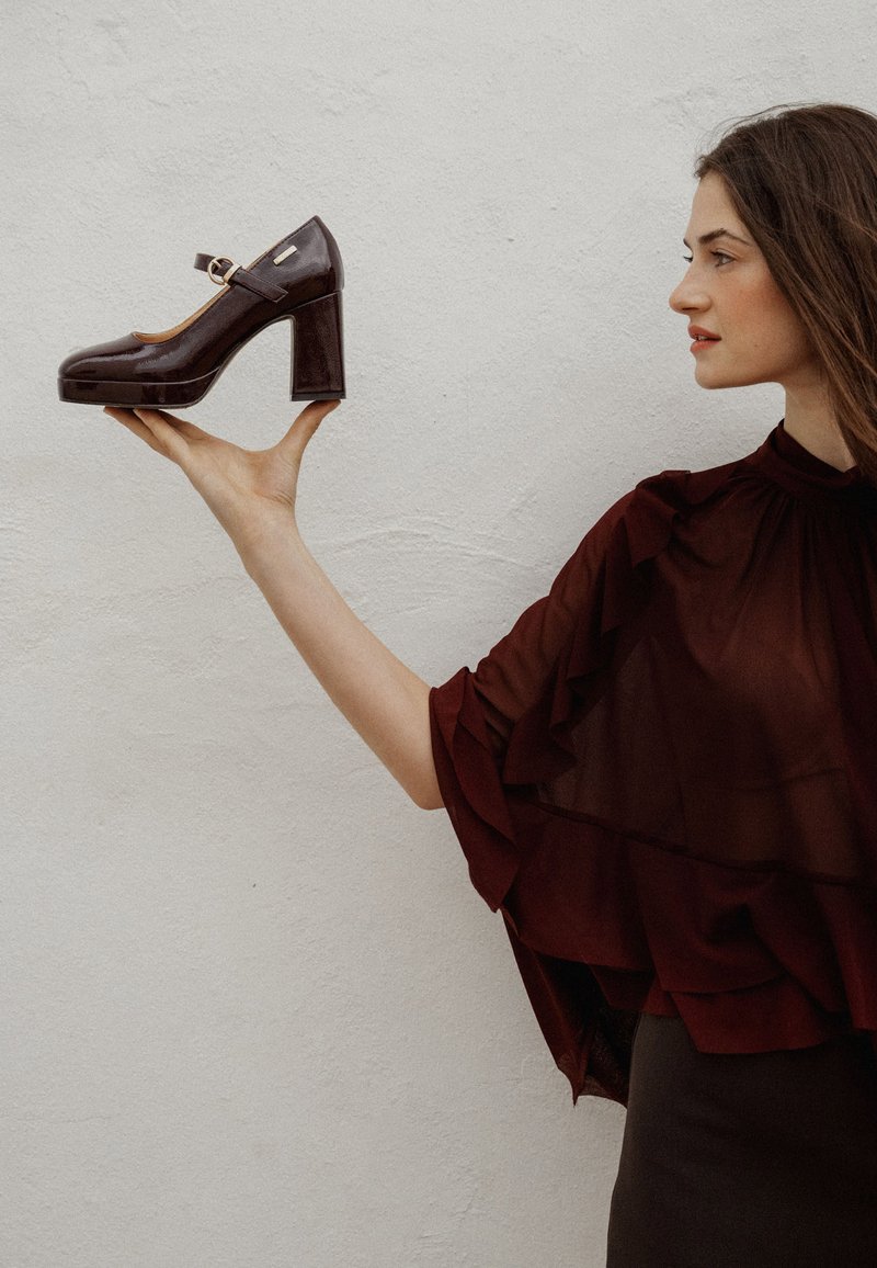 Chaussure à talon haut en cuir verni marron foncé avec un bout arrondi, une bride autour de la cheville et un talon épais, tenue devant un fond clair.