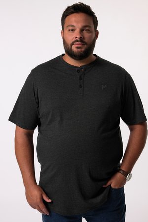Man with beard wearing dark gray short-sleeve henley shirt and wristwatch, standing with one hand in pocket against plain background.