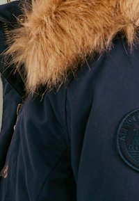 Navy blue jacket with a textured fur hood trim, featuring a round patch on the sleeve with an emblem, and a durable fabric design.