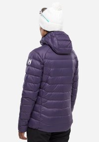 Doudoune violette avec capuche, présentant une texture matelassée et rembourrée. Inclus un logo sur l'épaule et un bonnet blanc avec un pompon.