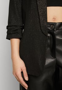 Vue recadrée d'une personne portant un blazer noir étincelant, un pantalon en cuir noir et un top court noir laissant apparaître un nombril visible.