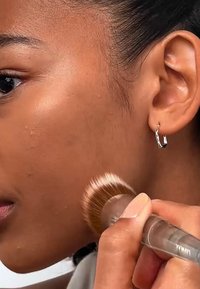 Makeup-børste med myke børstehår påfører foundation på huden. Modellen har glatt, solbrun hud og har på seg en enkel sølv hoop øredobb.