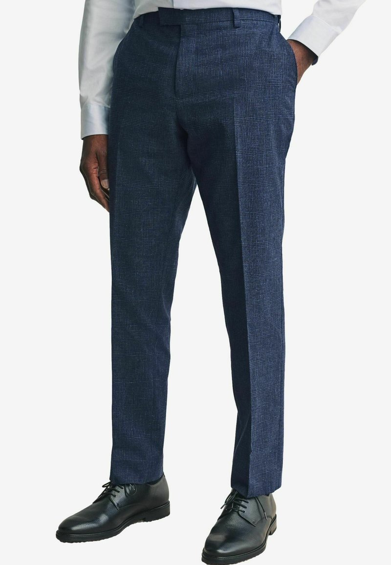 Marineblauwe herentrousers gemaakt van gestructureerde stof, met een recht gesneden ontwerp, een plooi aan de voorkant en zwarte nette schoenen.