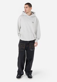 Felpa grigia chiara con cappuccio e tasca anteriore, abbinata a pantaloni neri larghi con cuciture a contrasto e un design di tasche unico. Sneakers nere.