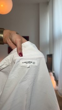 Witte katoenen shirt met een etiket waarop "PULL&BEAR" staat, maatdetails en "Gemaakt in India." Glad textuur, standaard kraag en lange mouwen.