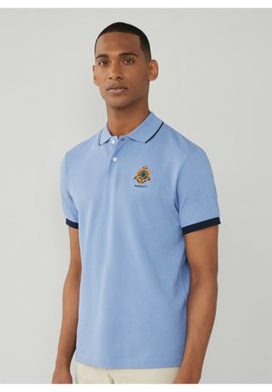 Camisa polo de color azul claro con mangas cortas, ribete marino en el cuello y los puños, con un logo bordado en el lado izquierdo del pecho. Textura suave.