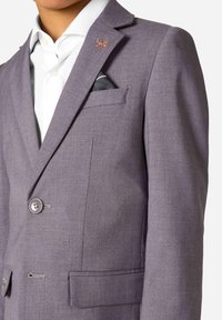 Dziecko w dopasowanej szarej marynarce z białą koszulą, chusteczką w kieszeni oraz pomarańczowym i srebrnym przypinką na klapie.