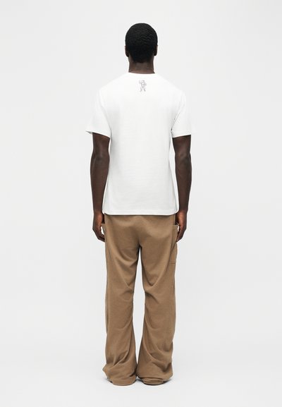 Άνδρας που στέκεται γυρισμένος με την πλάτη φορώντας λευκό κοντομάνικο μπλουζάκι με μικρό μαύρο γραφικό φιγούρας στο άνω μέρος της πλάτης και φαρδιά καφέ παντελόνια.