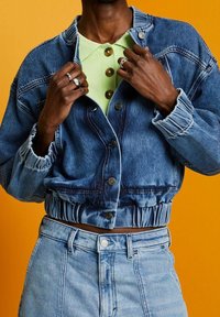 Jeansjacka med kort längd, knappstängning, elastisk fåll och fransade manschetter, bärs över en mintgrön skjorta med krage och bruna knappar.