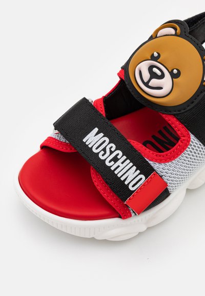Sandálias vermelhas e pretas com parte superior em malha, apresentando um detalhe em forma de rosto de urso. Solado branco espesso, tira ajustável com a marca "MOSCHINO". Textura suave.