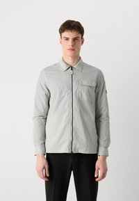 Φωτει�νό πράσινο μπουφάν με φερμουάρ σε υφή. Διαθέτει γιακά που διπλώνει, μπροστινή τσέπη και μακριά μανίκια. Μαύρες λεπτομέρειες στη μανσέτα.