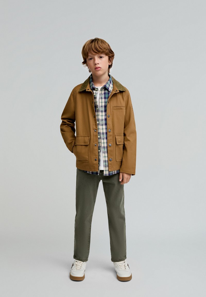 Chaqueta ligera marrón con un cuello verde, camisa de cuadros debajo, pantalones verde olivo y zapatillas blancas, de un niño que está de pie contra un fondo sencillo.