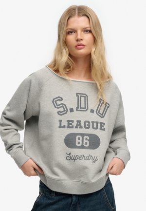Lyshåret kvinde iført en lysegrå sweatshirt med teksten "S.D.U LEAGUE 86 Superdry", poserer med hænderne i lommerne mod en hvid baggrund.