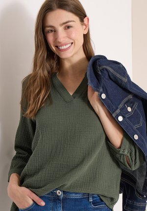 Femme souriante aux longs cheveux bruns portant un chemisier vert texturé et un jean bleu, tenant une veste en denim foncé pliée sur son épaule.
