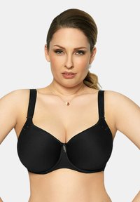 Donna che indossa un reggiseno nero con coppe sagomate, dotato di spalline regolabili e un piccolo charm decorativo al centro davanti, su uno sfondo semplice.