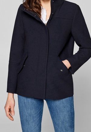 Frau trägt eine dunkel marineblaue Jacke mit Frontreißverschluss und Druckknöpfen, kombiniert mit blauen Jeans und einem weißen Hemd darunter.