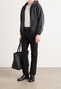 Schwarze Kapuzenjacke mit elastischen Bündchen, weißes T-Shirt mit Logo, schwarze Hose, schwarzer Gürtel und schwarze Schuhe. Trägt eine große schwarze Tragetasche.