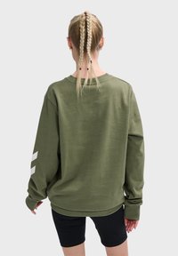 Persona con trenzas rubias vestida con una sudadera verde oliva de gran tamaño con rayas blancas en las mangas y pantalones cortos negros, de espaldas.