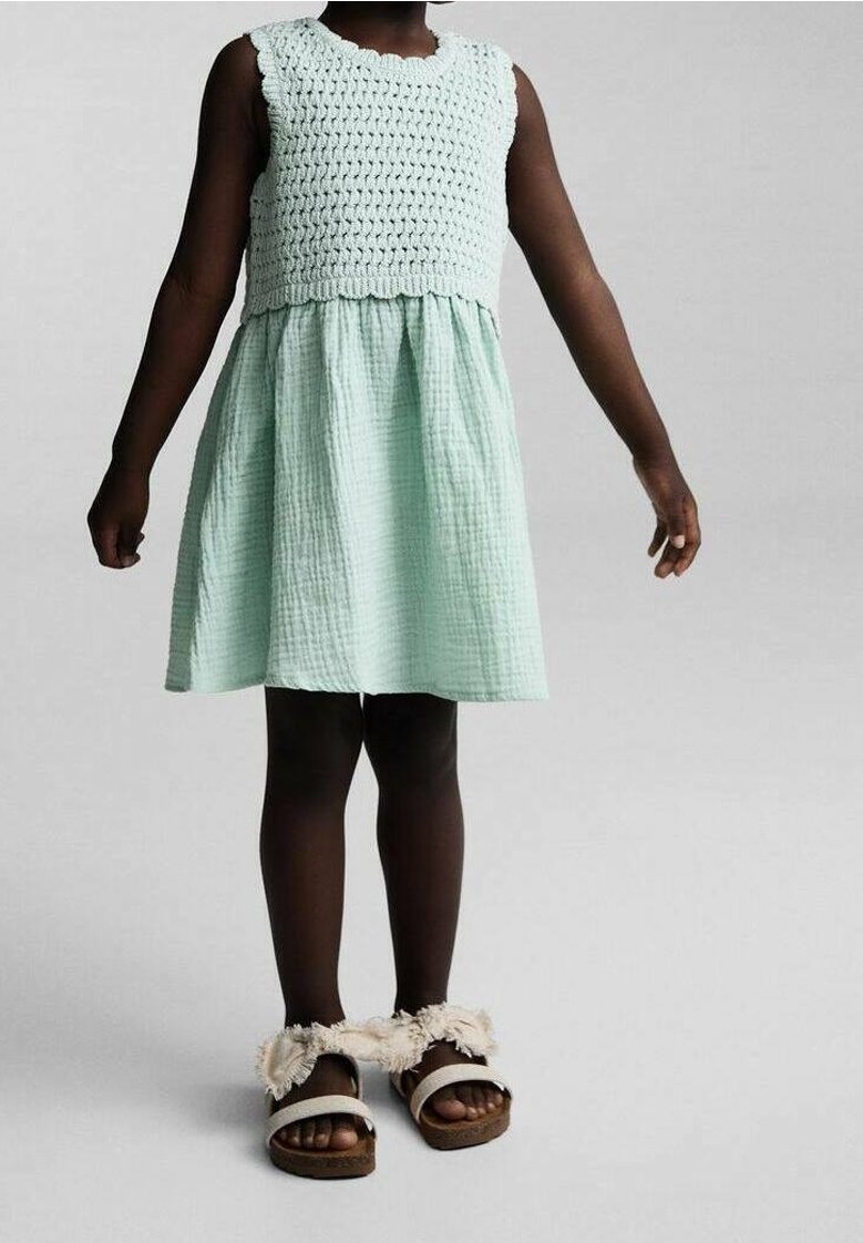 Enfant portant une robe sans manches vert menthe avec un haut texturé et des sandales à brides en fourrure beige, debout devant un fond uni.