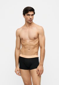 Homme torse nu aux cheveux courts et foncés, portant un boxer noir avec une ceinture orange clair portant la marque Lacoste, debout devant un fond blanc uni.