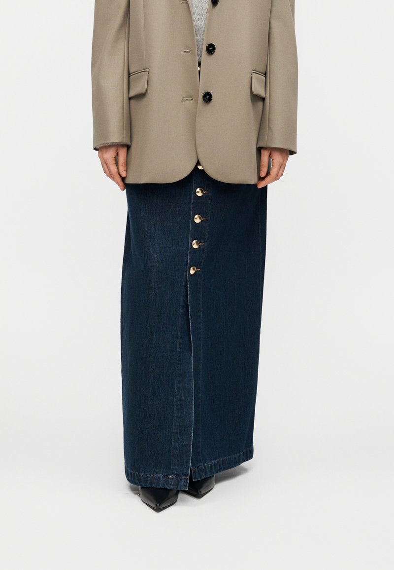 Μακριά φούστα από σκούρο τζιν με κλείσιμο με κουμπιά μπροστά, σε ίσια γραμμή. Συνδυασμένη με ανοιχτό γκρι τοπ και μπεζ oversized σακάκι.