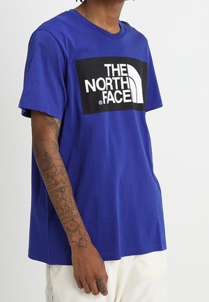 T-shirt bleu à manches courtes en coton, avec un grand rectangle noir contenant du texte blanc et un design de logo incurvé.