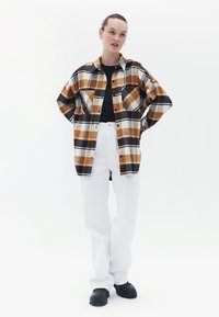 Camisa de cuadros en tonos marrón, negro y blanco, con dos bolsillos en el pecho y un corte relajado, combinada con pantalones anchos blancos y zapatos negros.