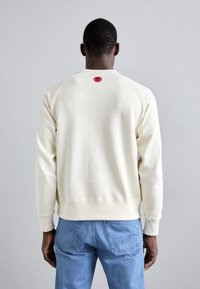 Felpa crema con scollo rotondo e maniche raglan, caratterizzata da una grafica di un sole rosso sul retro. Abbinata a jeans in denim blu.