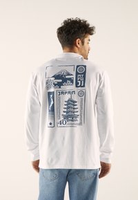 Langermet hvit T-skjorte med et blått grafisk motiv på baksiden, som viser Fuji-fjellet, en japansk pagode og elementer relatert til Japan.