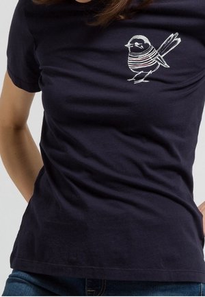 T-shirt bleu marine avec un petit oiseau blanc portant un pull rayé imprimé sur le côté gauche de la poitrine, porté par une personne les mains derrière le dos.