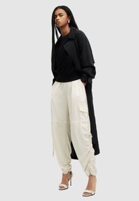 Zwarte lange jas over een zwart sweatshirt, met crèmekleurige cargo-broeken met zijzakken en aansnoerbare manchetten. Gestyled met open teen hakken.