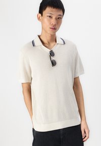 Nura trumpa rankovėmis polo marškiniai off-white spalvos, su tekstūruotu raštu ir tamsiai mėlynai dryžuotu apykakle. Modelis dėvi akinius nuo saulės.