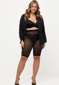Reggiseno nero e pantaloni corti con bordo in pizzo e motivo a diamante, indossati sotto una camicia nera aperta. La modella indossa tacchi neri, evidenziando le texture.