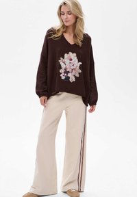 Brązowy oversize sweter z kwiatowym wzorem z przodu, w połączeniu z beżowymi szerokimi spodniami z bocznymi paskami w ciemnej czerwieni i bieli.