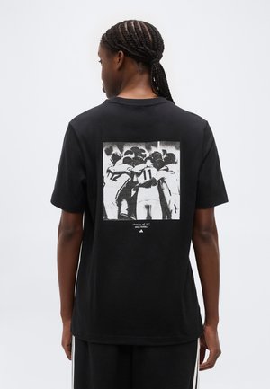 Põimitud juustega inimene, kes kannab musta t-särki, näitab mustvalget fotot jalgpallurite kokkukuuluvusest seljal.