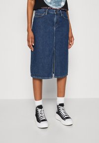 Džínová sukně ve tmavě modré barvě s předním rozparkem, zdobnými švy a klasickým střihem, kombinovaná s černými vysokými teniskami a bílými kotníkovými ponožkami.