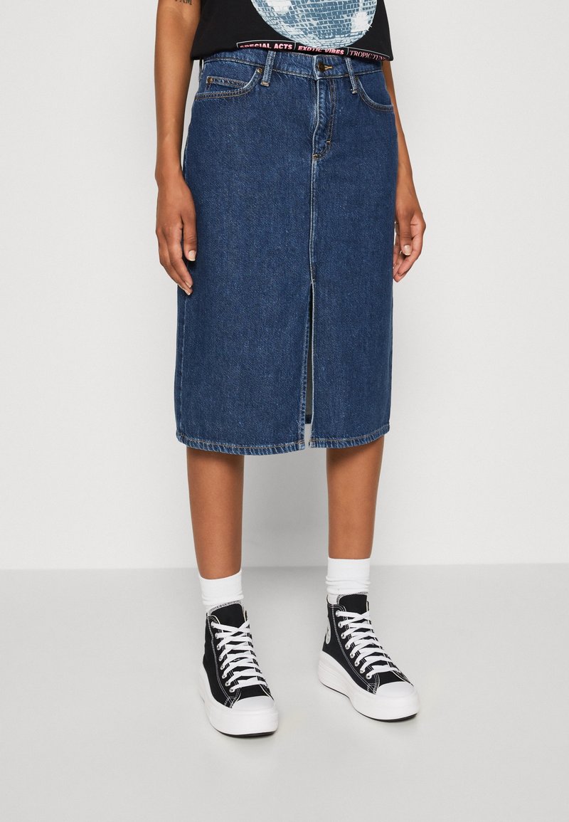 Džínová sukně ve tmavě modré barvě s předním rozparkem, zdobnými švy a klasickým střihem, kombinovaná s černými vysokými teniskami a bílými kotníkovými ponožkami.