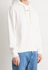 Personne portant un sweat à capuche blanc avec le texte « HUGO » sur la poitrine et un jean bleu clair, debout devant un fond blanc uni.