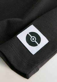 Mustaline musta tekstili, volditud äärega, millel on valge silt musta ringi kujutisega Pokémoni logoga. Sile tekstuur, minimalistlik disain.