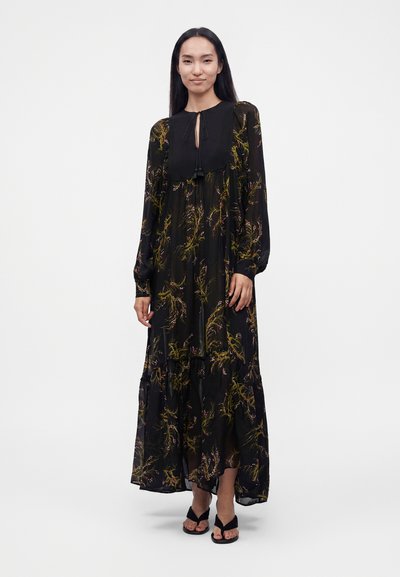 Femme aux longs cheveux foncés porte une robe maxi noire à fleurs avec un décolleté en goutte d'eau et des manches longues, debout sur un fond blanc uni.