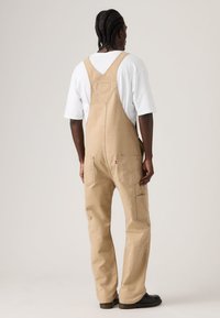 Άνδρας με πλεξούδες, φοράει μπεζ ολόσωμη φόρμα και άνετο λευκό t-shirt, στέκεται με την πλάτη προς το μέρος του θεατή σε απλό φωτεινό φόντο.