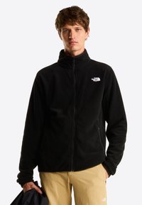 Ung mand iført en sort North Face fleecejakke og beige bukser, der holder et sort stykke tøj i sin venstre hånd.