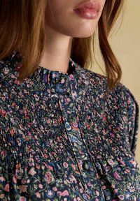 Sieviete, kura valkā tumšu ziedu daudzslāņu blūzi ar augstu frillētu apkakli un garām piedurknēm, ar gaiši brūniem matiem, kas daļēji redzami.