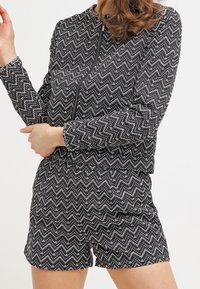 Frau in einer Jacke mit langem Ärmel im schwarz-weißen Zickzackmuster und passenden hochgeschnittenen Shorts, stehend mit verschränkten Armen.