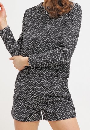 Vrouw in een zwart-wit zigzagpatroon jas met lange mouwen en bijpassende highwaisted shorts, staand met gekruiste armen.